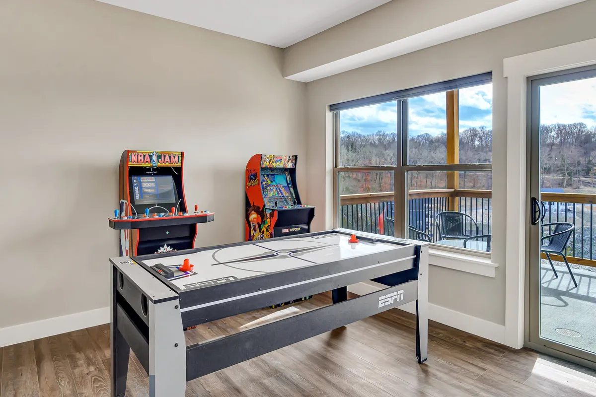 A vibrant game room featuring an air hockey table in the foreground, several classic arcade machines along the wall, and large windows that provide scenic views. The space feels playful and energetic, inviting guests of all ages to enjoy friendly competition and entertainment.