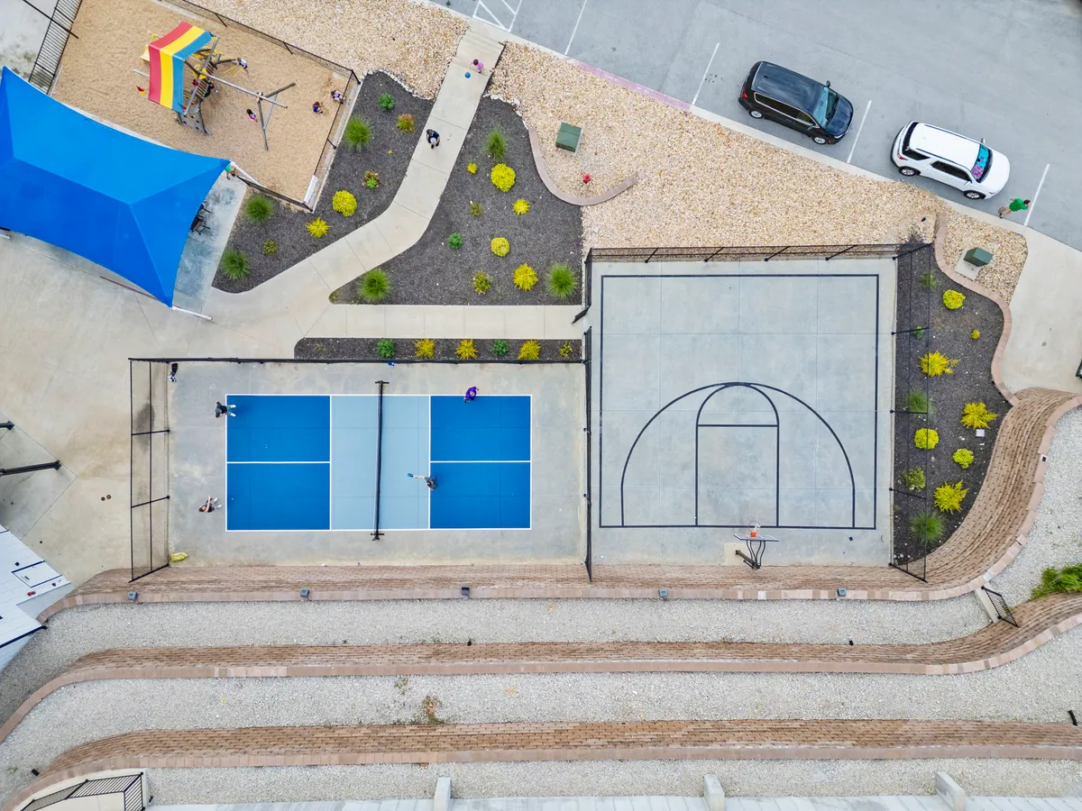A lively exterior view capturing tennis courts, a basketball court, and a playground area, all surrounded by landscaping. The space shows multiple recreational options with people enjoying games and socializing.