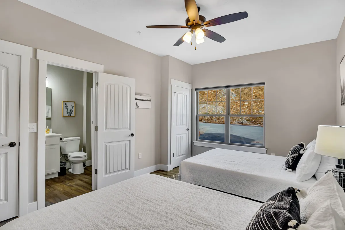 This image shows a cozy bedroom featuring two beds positioned near a window, with natural light streaming in. An ensuite bathroom is visible, highlighting the convenience and privacy of the space. The decor is simple and calming, enhanced by soft linens and a neutral palette.