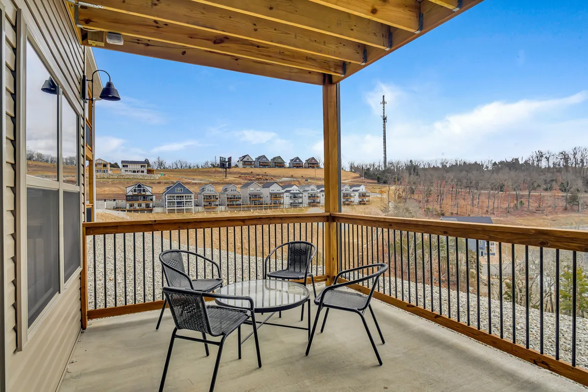 The image showcases a wooden porch with metal chairs and a glass table, set against a backdrop of rolling hills and neighboring cabins. The setting suggests enjoying morning coffee or leisurely conversations while immersed in peaceful natural surroundings.