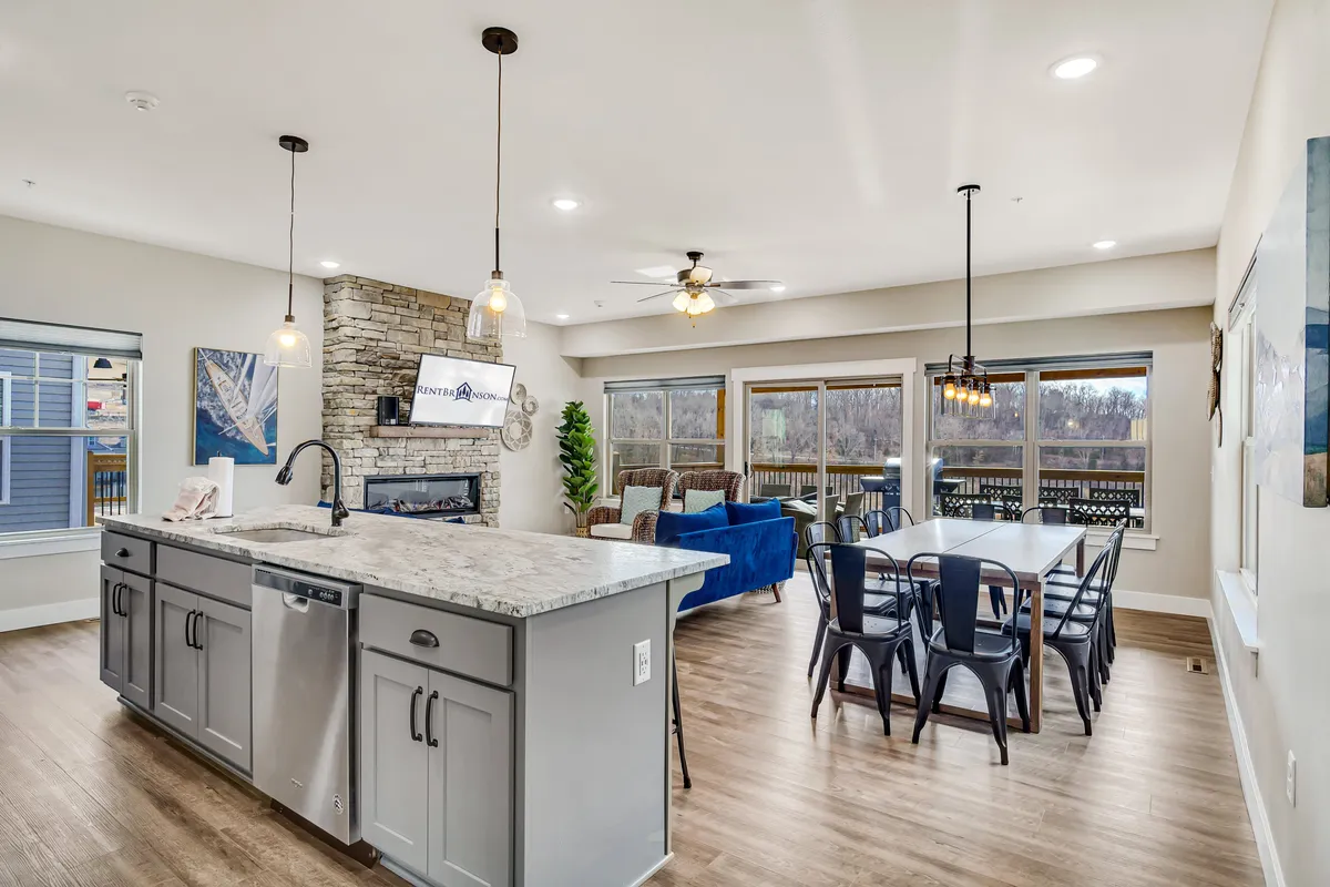 An open-concept space with a kitchen island, stone accent wall, dining table, and living area seating. The kitchen is modern and fully equipped, while the dining and living areas flow seamlessly together, framed by windows that let in natural light.