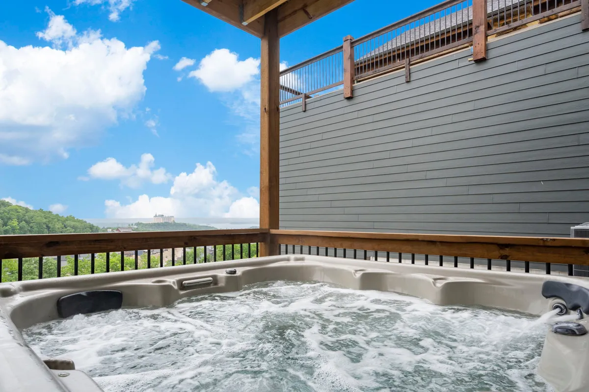 An inviting outdoor hot tub occupies the foreground, overlooking a beautiful natural landscape with Table Rock Lake in the distance under a cloudy sky. The serene vibe offers a perfect contrast to the high-energy game lounge inside.