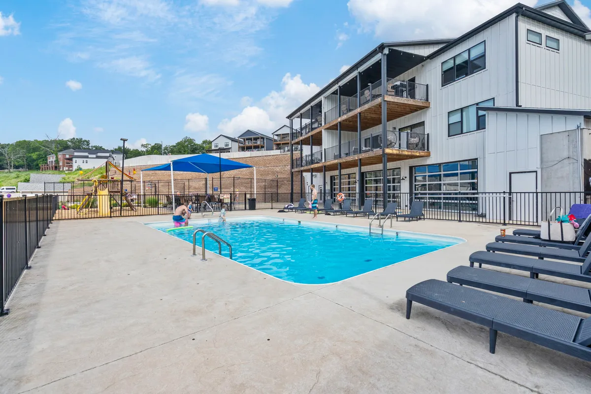 A vibrant exterior shot of the community's resort-style amenities, including a sparkling outdoor pool with lounge chairs, a colorful playground structure, and part of the building facade. Families and children can be seen enjoying the space.