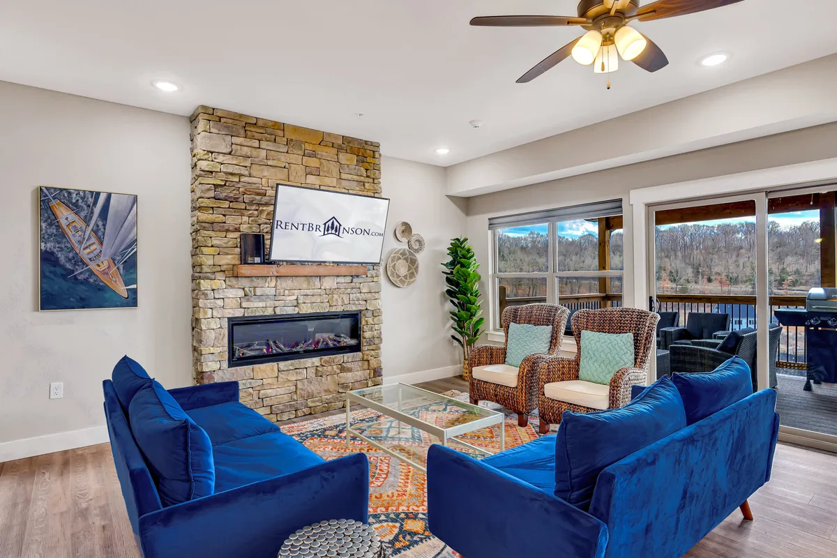 A welcoming living room featuring blue velvet sofas arranged around a stone fireplace, with a large flat-screen TV mounted above. The space is anchored by a colorful area rug, and sliding glass doors open out to a deck, letting in abundant natural light.