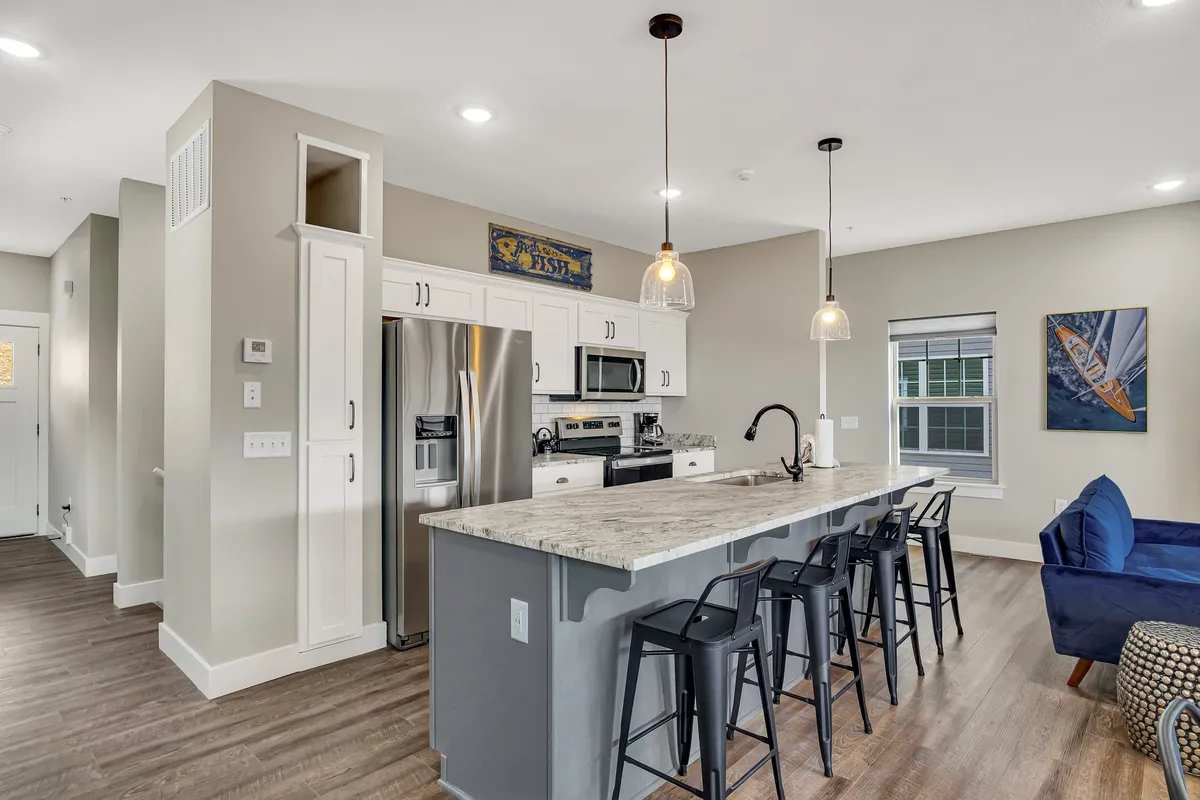 A stylish kitchen with a large island and bar stools, stainless steel appliances, and modern light fixtures. The kitchen opens into a living space with a blue couch and vibrant wall art, creating an inviting setting for cooking and relaxing.