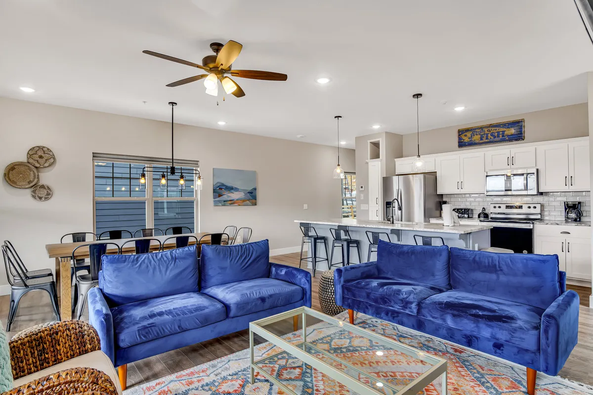A bright, spacious open-concept living area with plush blue sofas set around a coffee table, a modern dining table ready for shared meals, and a sleek kitchen island—inviting guests to gather, relax, and connect.